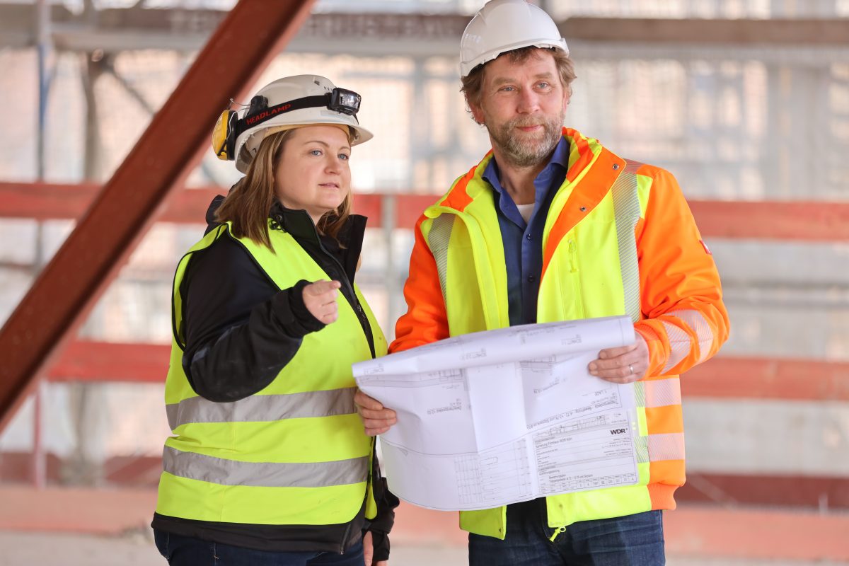 Eine Bauarbeiterin und ein Bauarbeiter prüfen Baupläne auf einer Baustelle. Beide tragen Schutzhelme und Warnwesten.